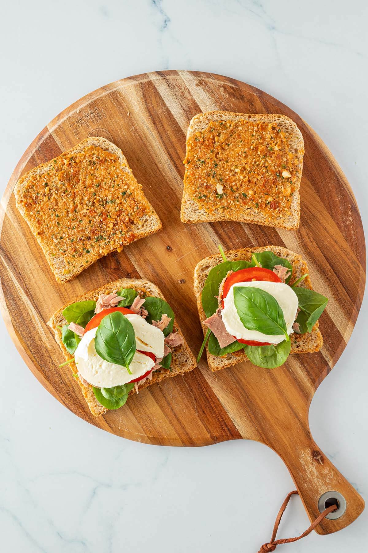 Vier mit Pesto bestrichene Toastscheiben auf einem Holzbrett, zwei davon mit Salat, Thunfisch, Tomate und Mozzarella belegt.