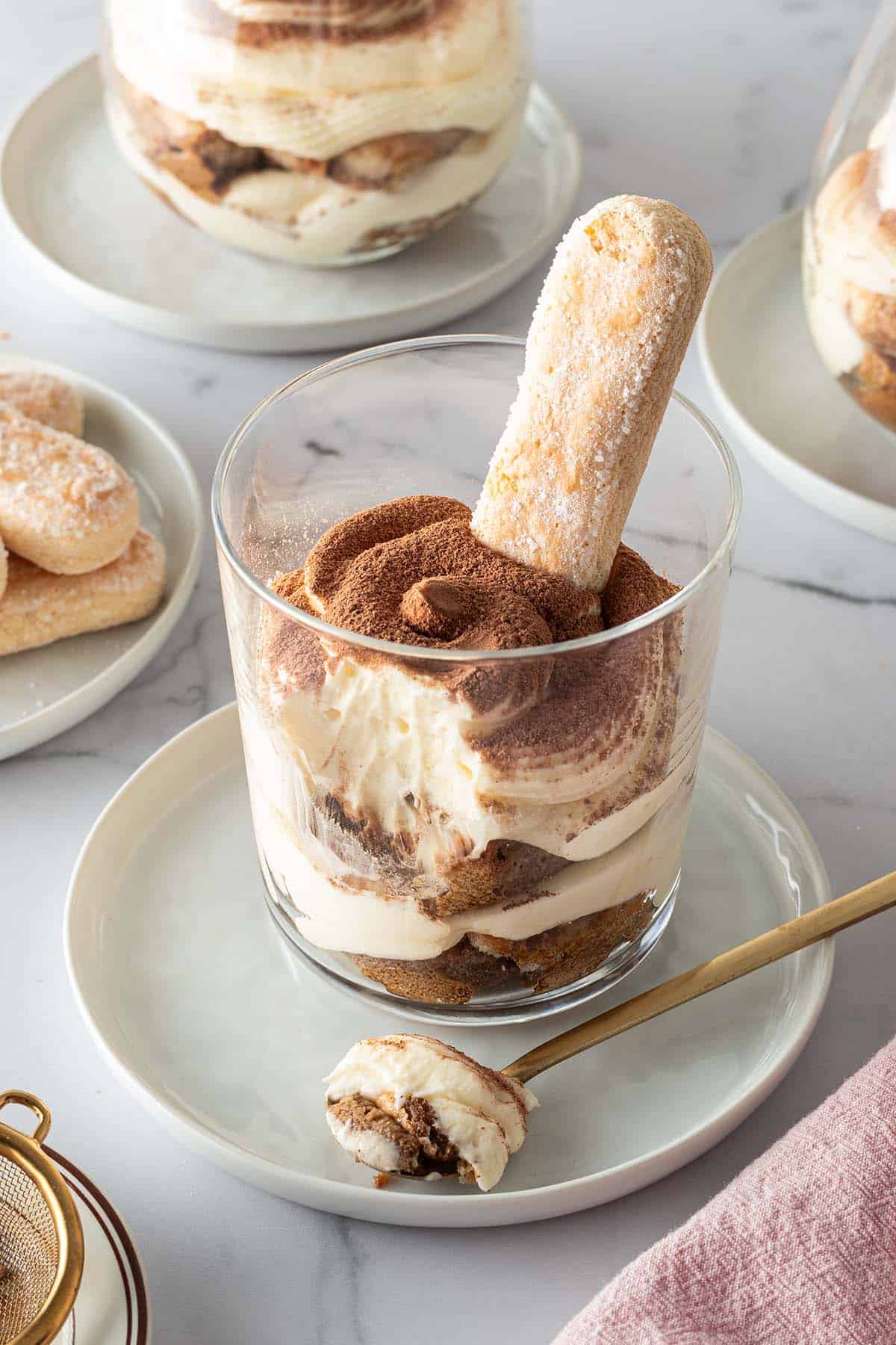 Tiramisu im Glas geschichtet mit L&ouml;ffelbiskuit, im Hintergrund mehr Gl&auml;ser mit Tiramisu und L&ouml;ffelbiskuits.