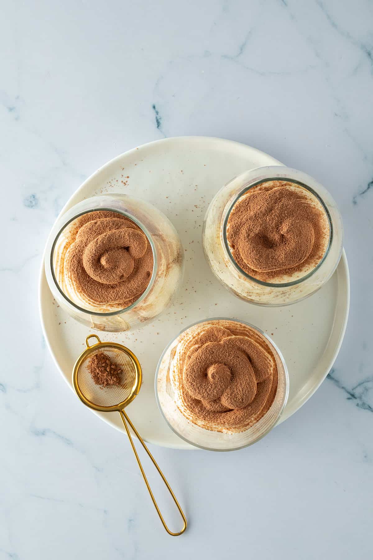 Drei Gl&auml;ser Tiramisu von oben, best&auml;ubt mit Kakao, daneben ein kleines Sieb.