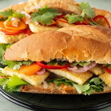 Zwei Tofu Banh Mi Sandwiches mit eingelegtem Gem&uuml;se und frischem Koriander auf einem Teller, Koriander und ein Glas im Hintergrund.