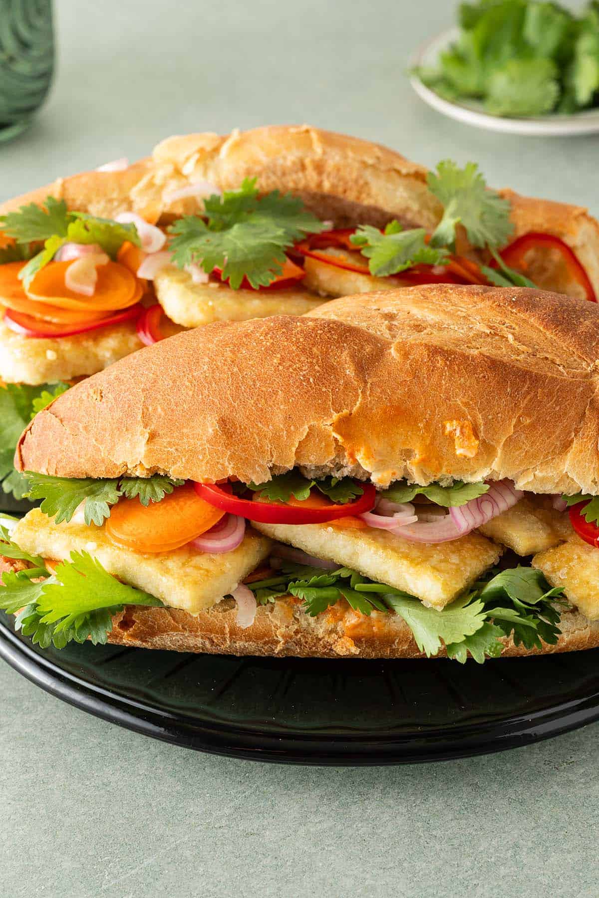 Zwei Tofu Banh Mi Sandwiches mit eingelegtem Gem&uuml;se und frischem Koriander auf einem Teller, Koriander und ein Glas im Hintergrund.