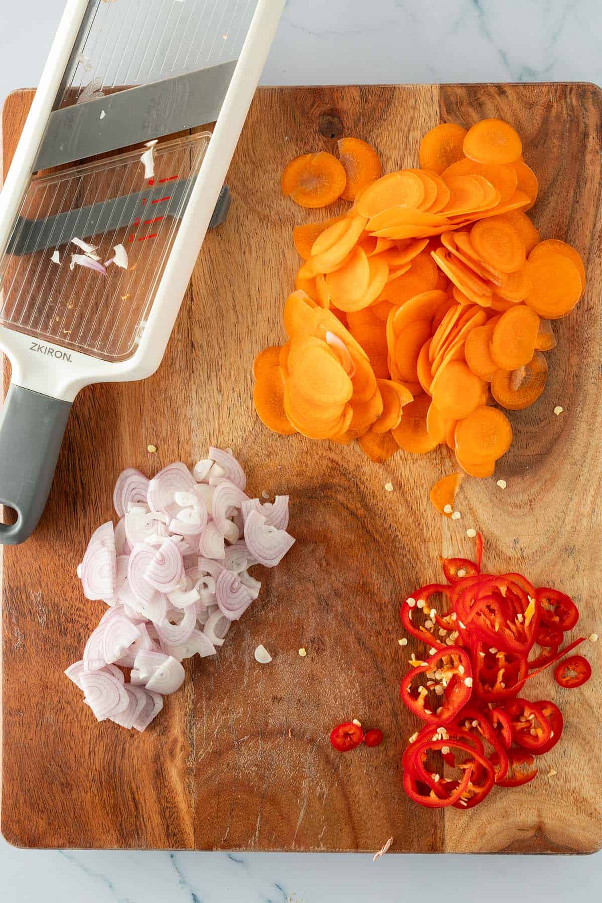In d&uuml;nne Scheibchen geschnittene Karotten, Schalotten und rote Chili auf Holzbrett mit einer Gem&uuml;sehobel daneben.