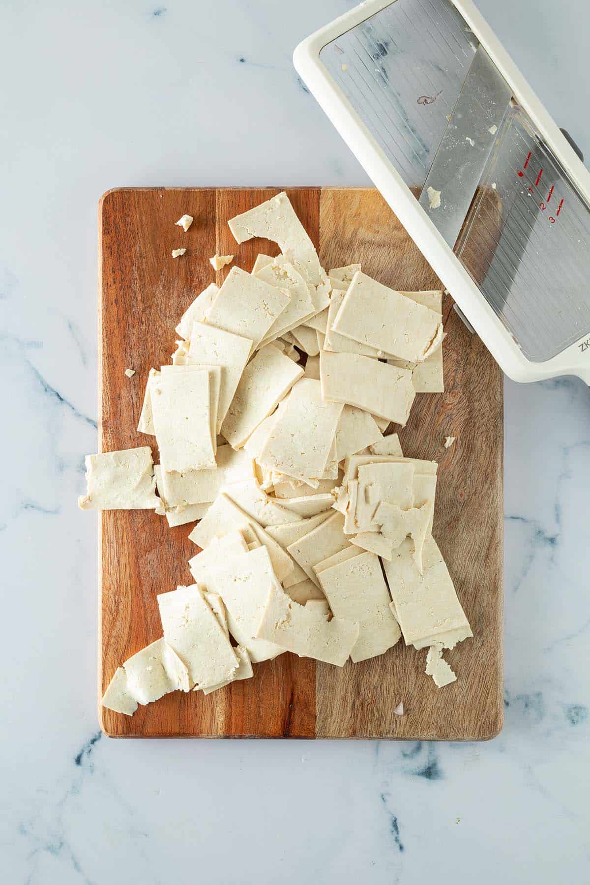 Sehr d&uuml;nne Tofu-Scheibchen auf einem Holzbrett, daneben eine Gem&uuml;sehobel.