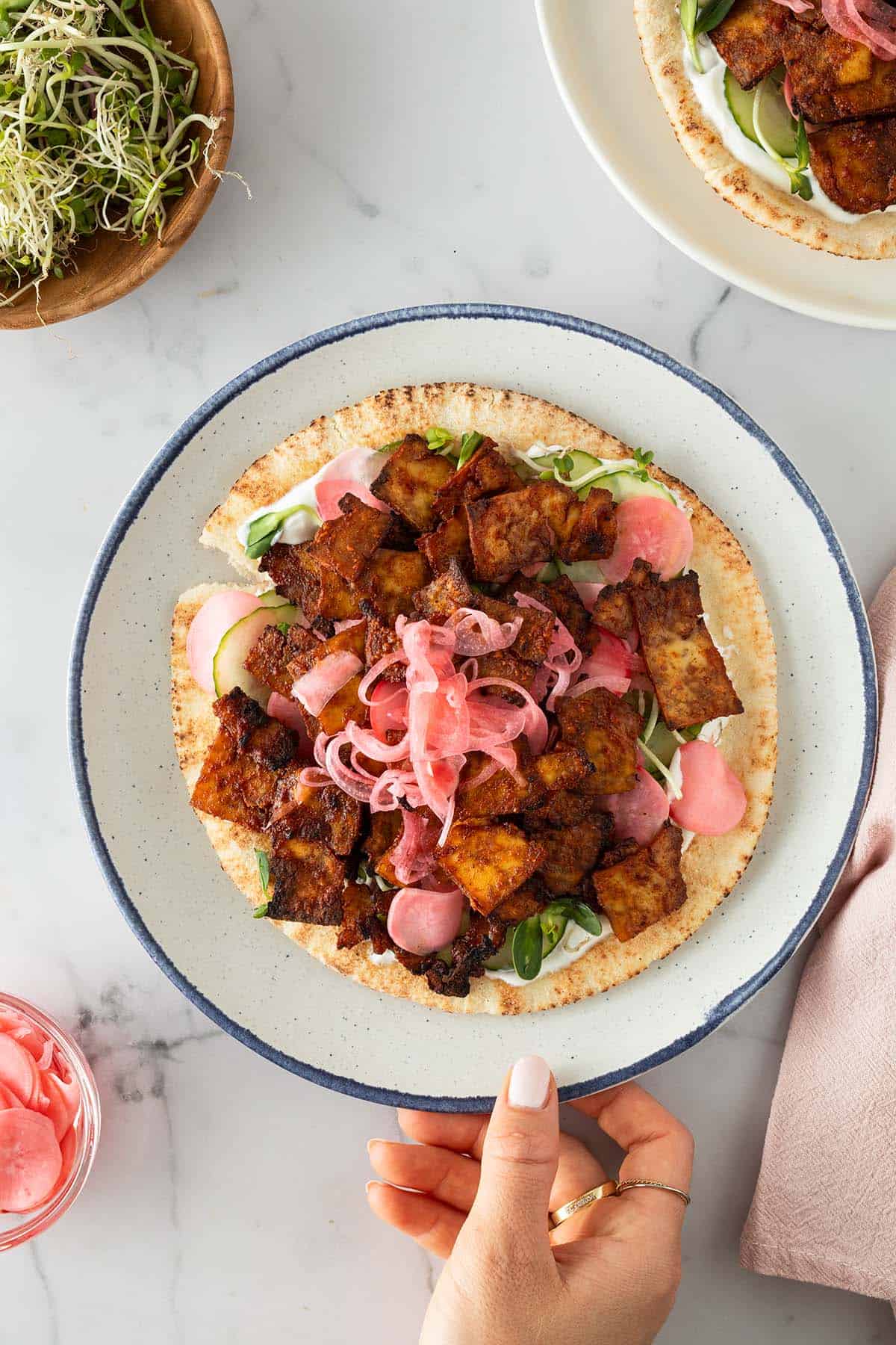 Fladenbrot belegt mit gebratenem Tofu-Gyros, Joghurtso&szlig;e, Gurkenscheiben, Sprossen und eingelegten roten Zwiebeln, serviert auf einem hellen Teller. Zutaten im Bildrand.