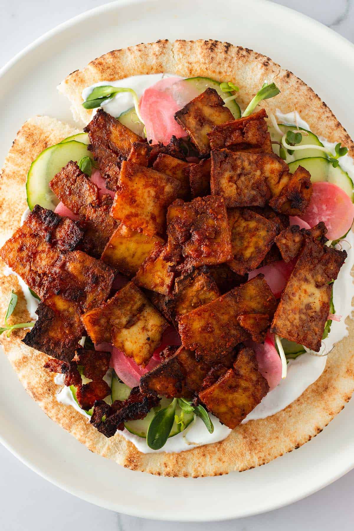 Nahaufnahme eines Pita-Brots getoppt mit knusprigem Tofu-Gyros, Gurkenscheiben, Sprossen, Joghurtso&szlig;e und eingelegten Zwiebeln.