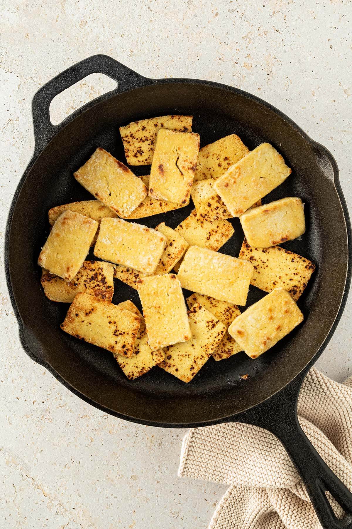 Knusprig gebratener Tofu in einer schwarzen Pfanne auf einem beigen Untergrund.