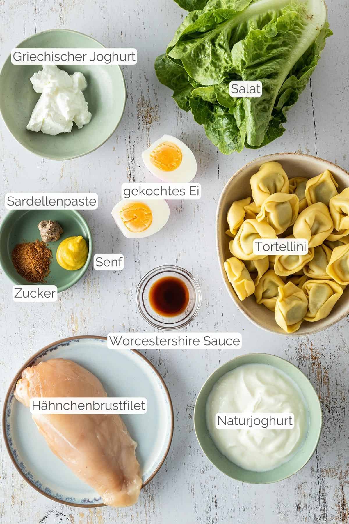 Alle Zutaten die für den Tortellini Salat ohne Mayo benötigt werden mit jeweiligen Textbeschriftungen.