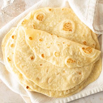 Ein Stapel selbstgemachte Weizen Tortillas auf einem hellen Tuch, mit Mehl und Limetten im Bildrand.