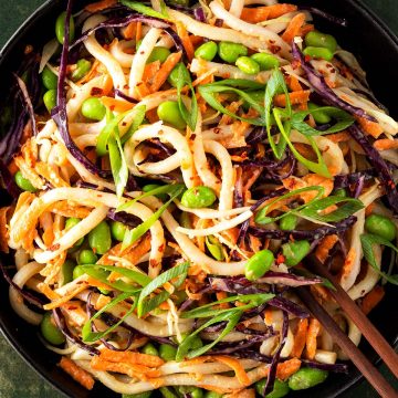 Udon Nudelsalat mit frischem Gem&uuml;se, Edamame Bohnen und einem Erdnussdressing in einem tiefen, schwarzen Teller.