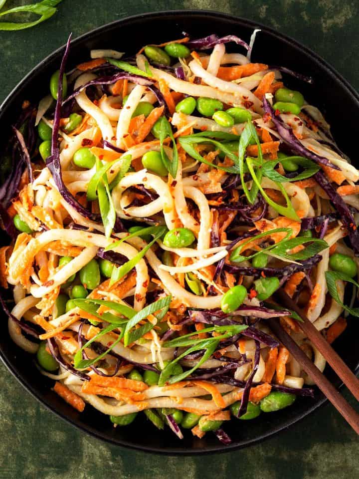 Udon Nudelsalat mit frischem Gem&uuml;se, Edamame Bohnen und einem Erdnussdressing in einem tiefen, schwarzen Teller.