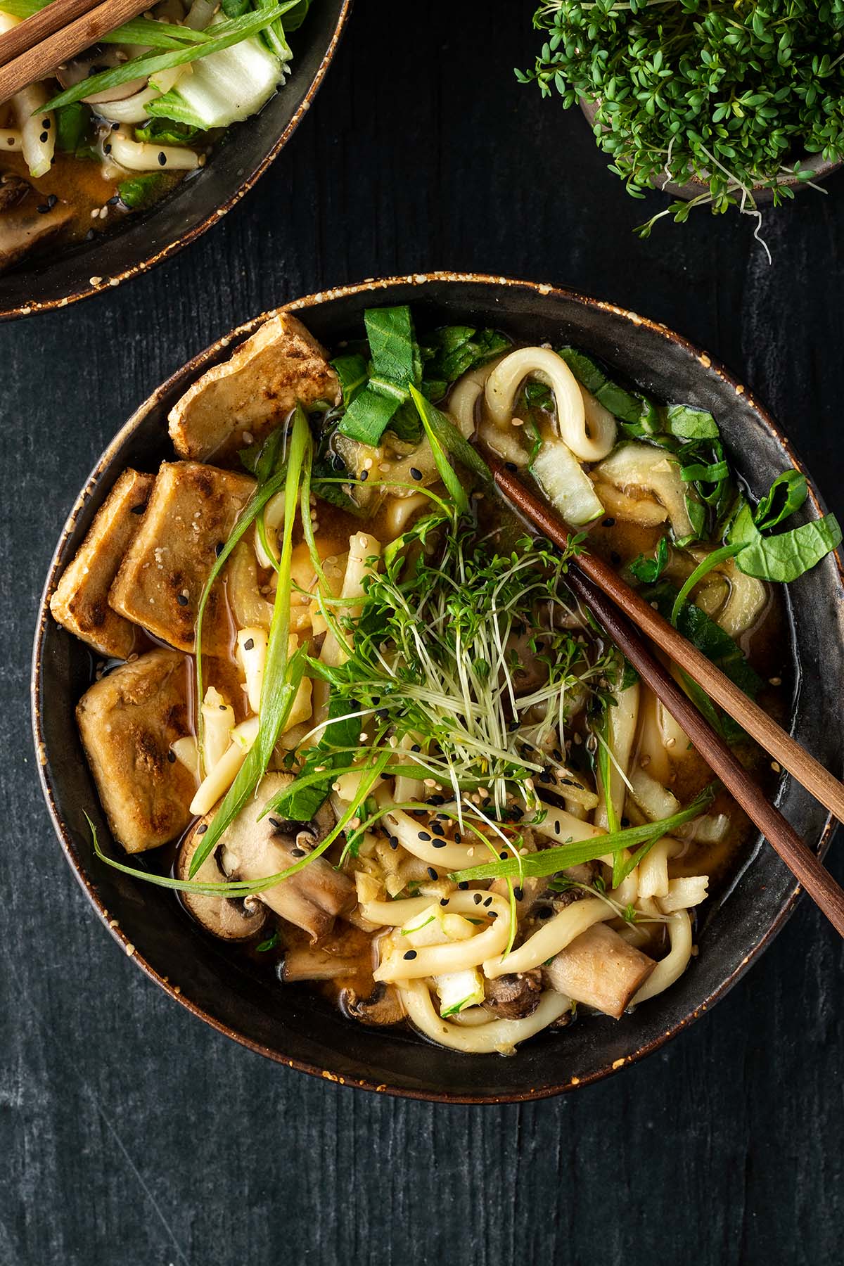 Eine Sch&uuml;ssel mit einer asiatischen Suppe mit dicken Udon Nudeln, Tofu, Pilzen und Gr&uuml;n auf einem dunklen Holztisch. Im Bildrand eine zweite Sch&uuml;ssel und eine Sch&uuml;ssel mit Kresse.