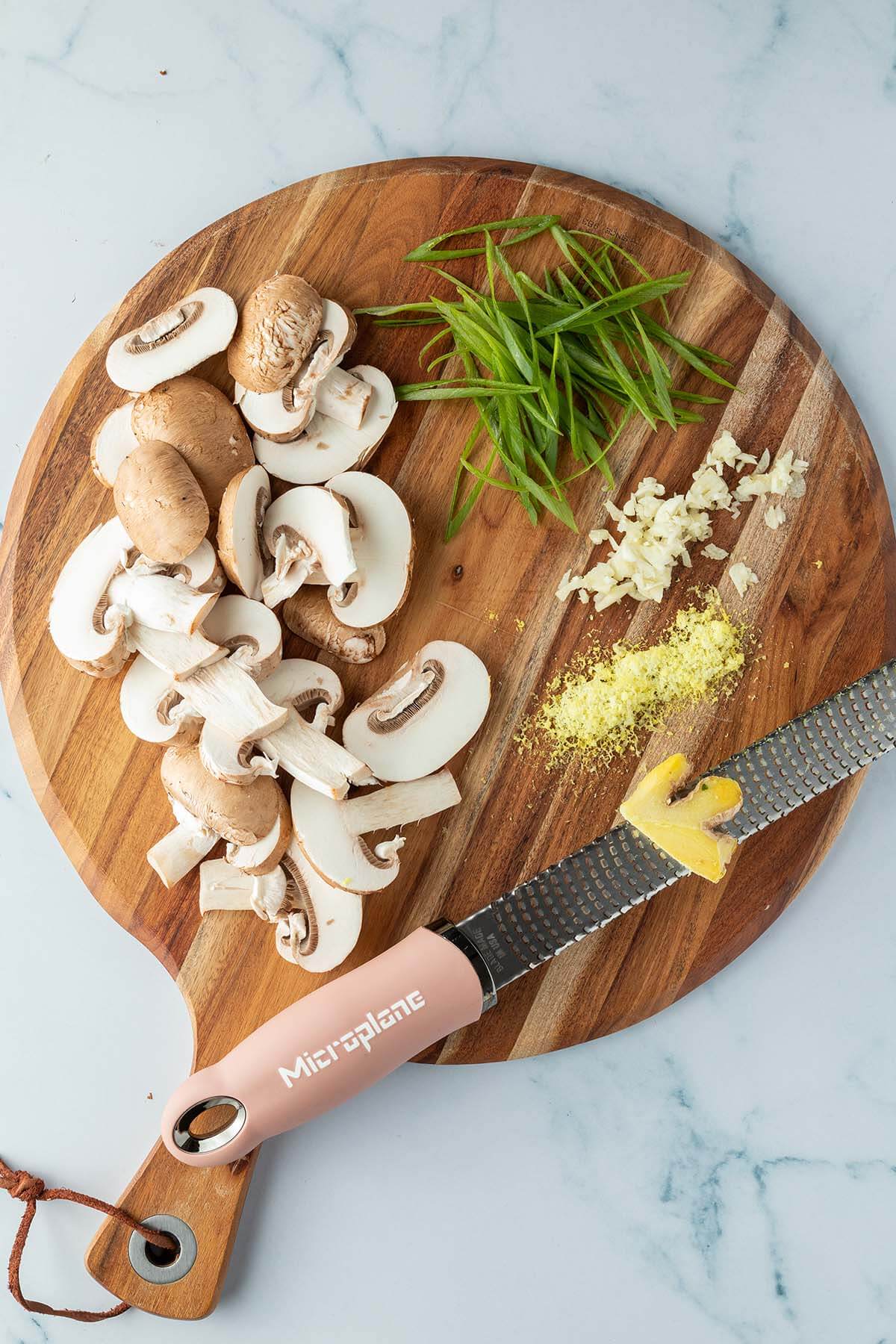 Ein Holzbrett mit in Scheiben geschnittenen Champignons, in d&uuml;nne Streifen geschnittenen Fr&uuml;hlingszwiebeln, fein gew&uuml;rfeltem Knoblauch und auf einer Microplane geriebenem Ingwer.
