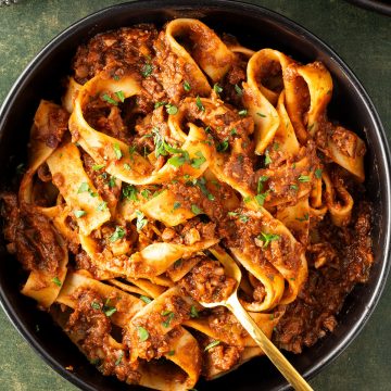 Ein Teller mit dicken, langen Nudeln in einer vegetarischen Bolognese auf dunkelgr&uuml;nen Untergrund, Weinglas und Teller im Bildrand.