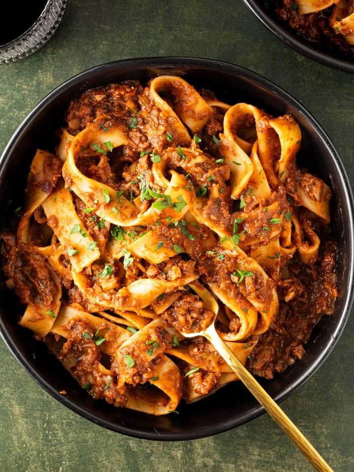 Ein Teller mit dicken, langen Nudeln in einer vegetarischen Bolognese auf dunkelgr&uuml;nen Untergrund, Weinglas und Teller im Bildrand.
