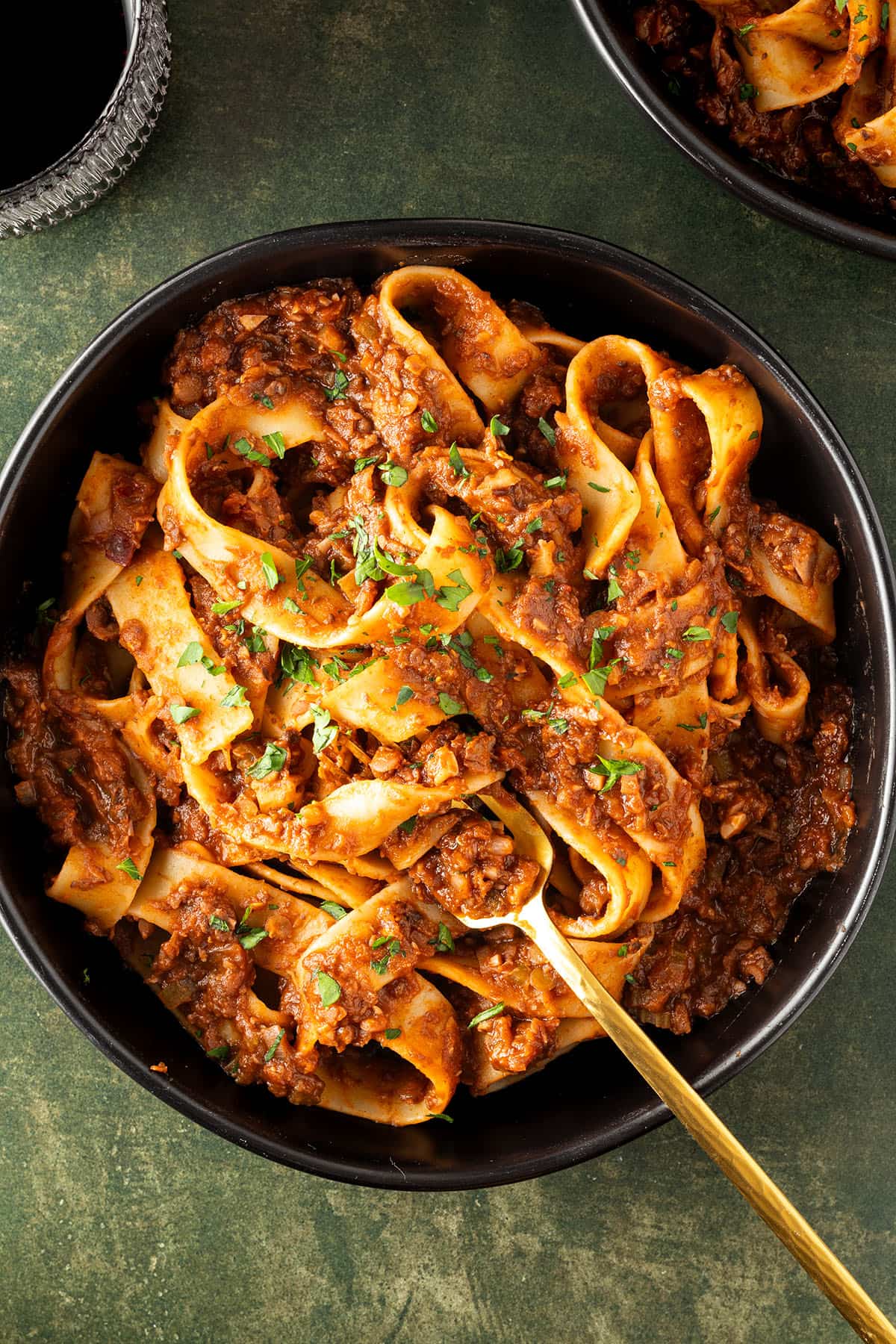Ein Teller mit dicken, langen Nudeln in einer vegetarischen Bolognese auf dunkelgr&uuml;nen Untergrund, Weinglas und Teller im Bildrand.