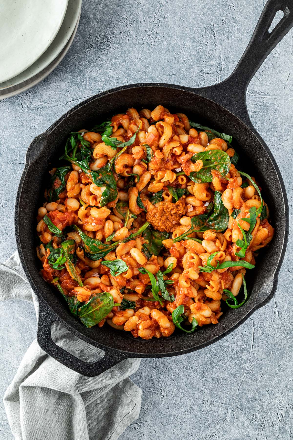 Vegetarische One Pot Pasta wird in einer Gusseisenpfanne serviert. Ein Tellerstapel im oberen Bildrand.