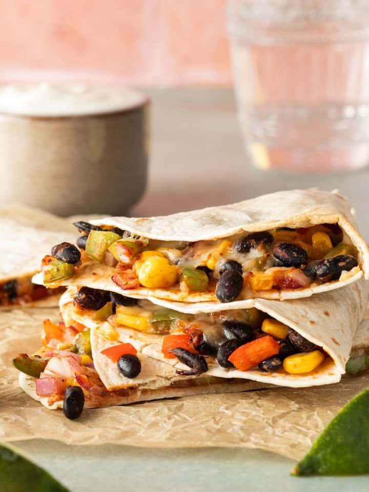 Ein Stapel aus vegetarisch gef&uuml;llten Quesadillas auf einem St&uuml;ck Backpapier mit Limettenspalten und einem Dip im Hintergrund.