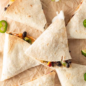 In Dreiecke geschnittene Quesadillas mit K&auml;se-Bohnen-Gem&uuml;se-F&uuml;llung auf einem Backpapier mit Jalape&ntilde;os und Limetten.