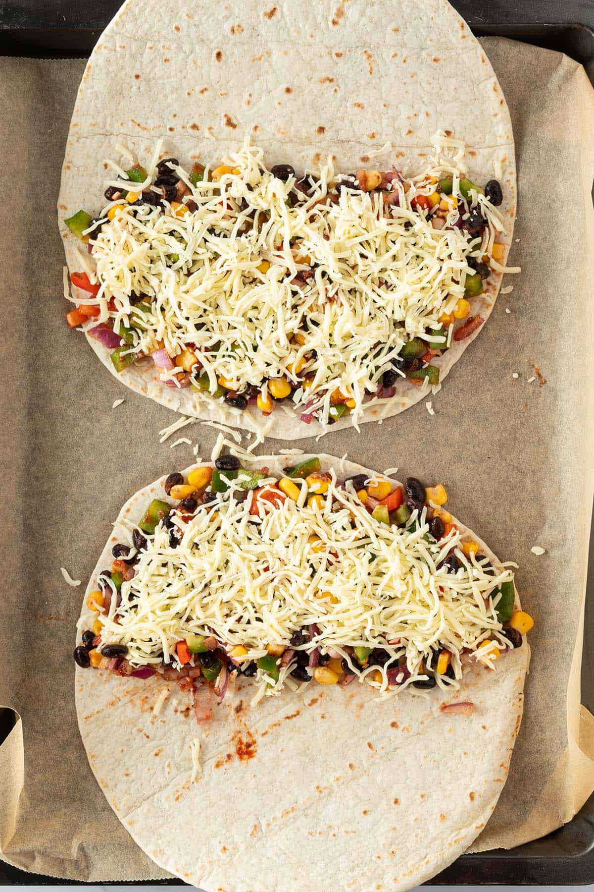 Zwei Tortillas auf einem Blech, die je zur H&auml;lfte mit der Gem&uuml;sef&uuml;llung und K&auml;se belegt sind.