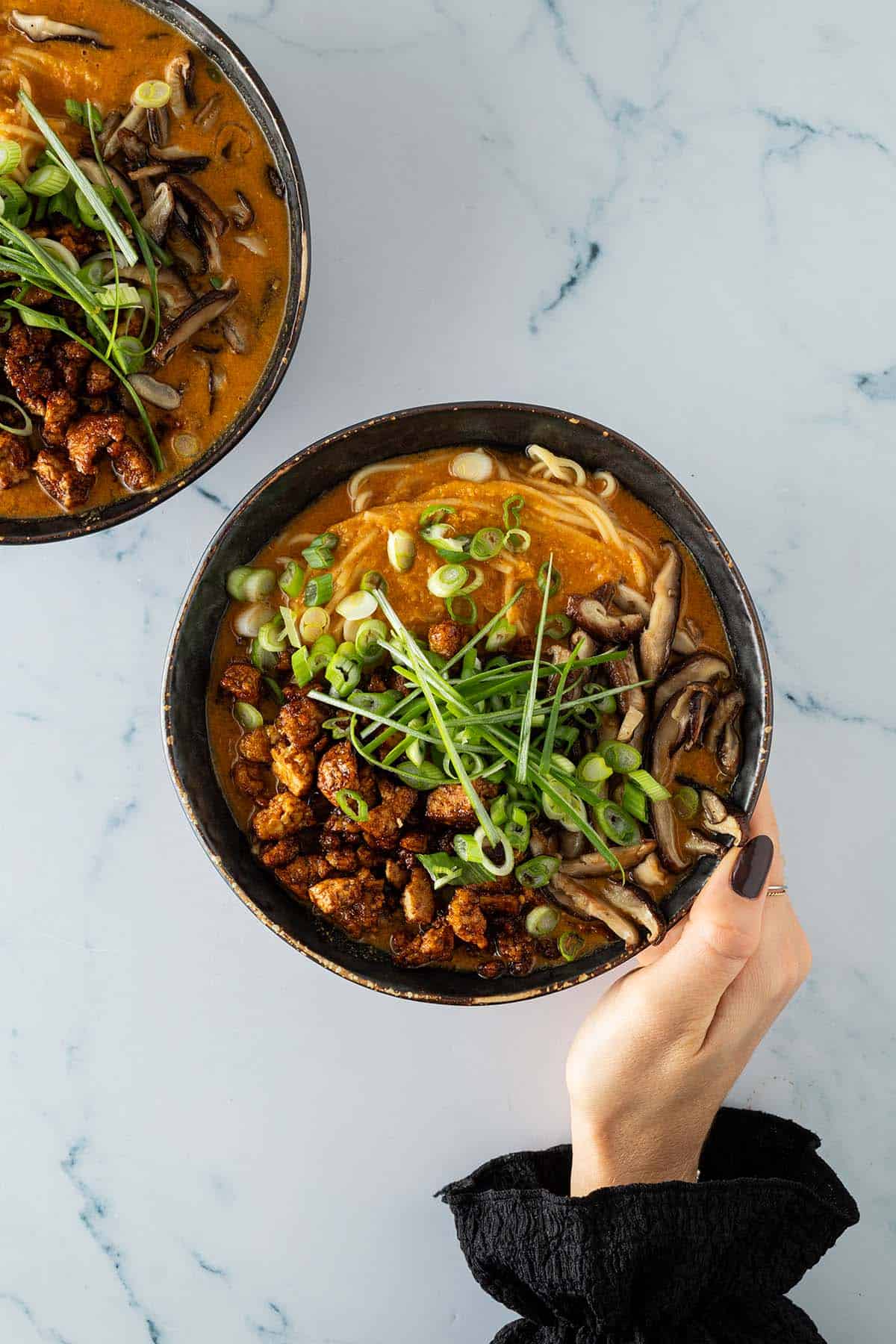 Zwei Sch&uuml;sseln vegetarische Erdnuss-Ramen auf heller Arbeitsfl&auml;che, serviert mit knusprigem Tofu, Pilzen und Fr&uuml;hlingszwiebeln.