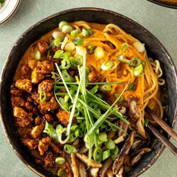 Cremige vegetarische Erdnuss-Ramen mit Ramennudeln, knusprig gebratenem Tofu, Shiitakepilzen und Fr&uuml;hlingszwiebeln in einer dunklen Sch&uuml;ssel von oben fotografiert.