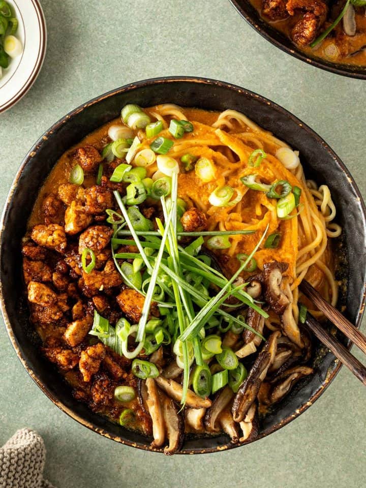 Cremige vegetarische Erdnuss-Ramen mit Ramennudeln, knusprig gebratenem Tofu, Shiitakepilzen und Fr&uuml;hlingszwiebeln in einer dunklen Sch&uuml;ssel von oben fotografiert.