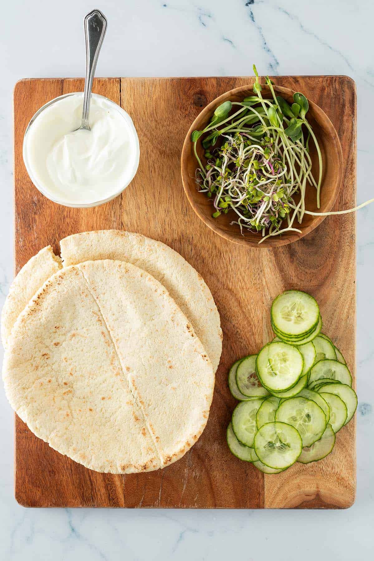Zutaten f&uuml;r den D&ouml;ner: zwei Pita-Fladenbrote, Gurkenscheiben, Sprossen und eine Schale mit Joghurtso&szlig;e auf einem Holzbrett angerichtet.