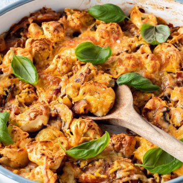Tortellini Auflauf in einer runden Auflaufform mit frischem Basilikum. Mit einem Holzl&ouml;ffel wird gerade eine Portion rausgenommen.