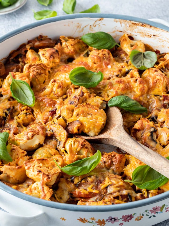 Tortellini Auflauf in einer runden Auflaufform mit frischem Basilikum. Mit einem Holzl&ouml;ffel wird gerade eine Portion rausgenommen.