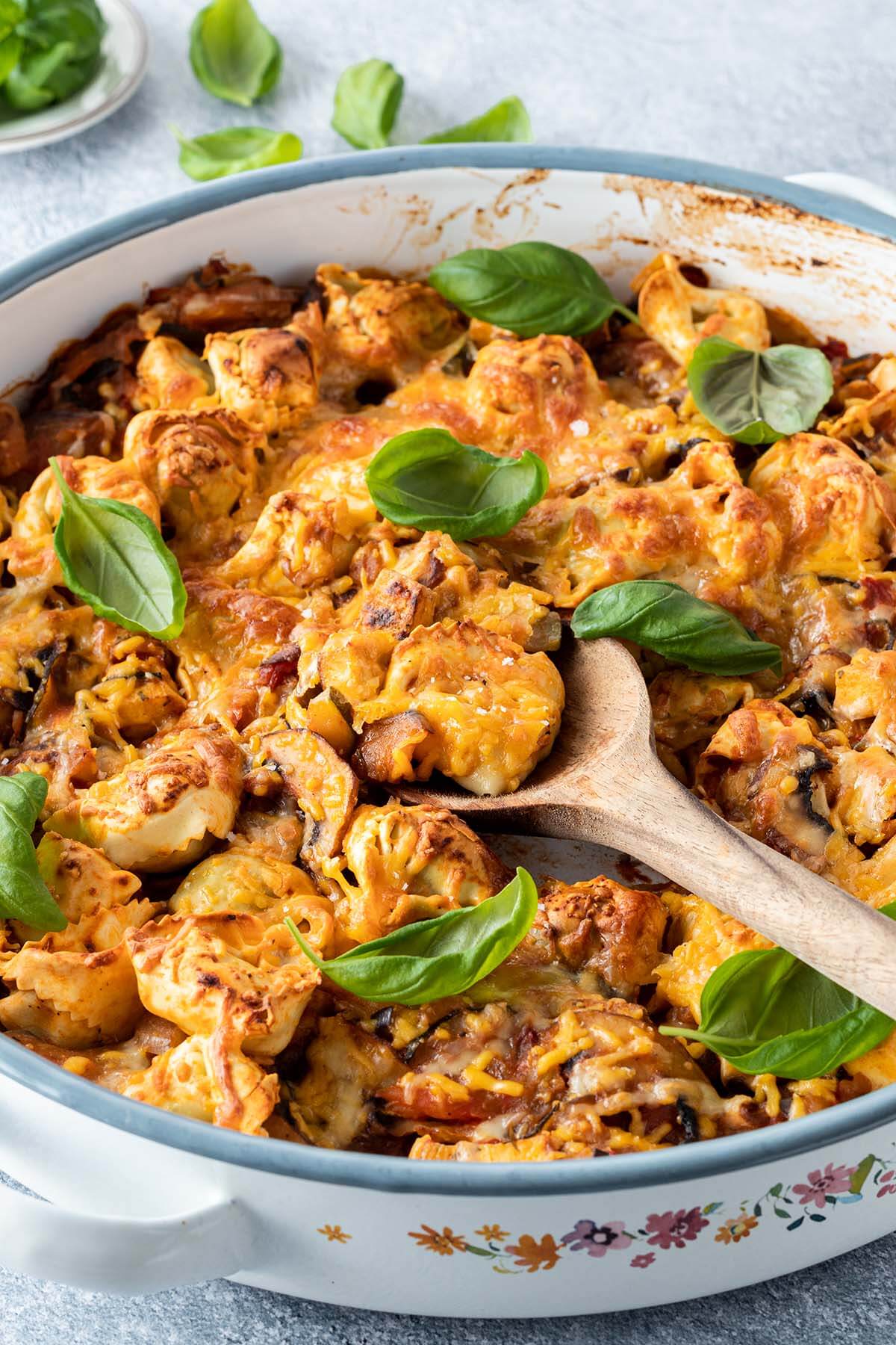 Tortellini Auflauf in einer runden Auflaufform mit frischem Basilikum. Mit einem Holzl&ouml;ffel wird gerade eine Portion rausgenommen.