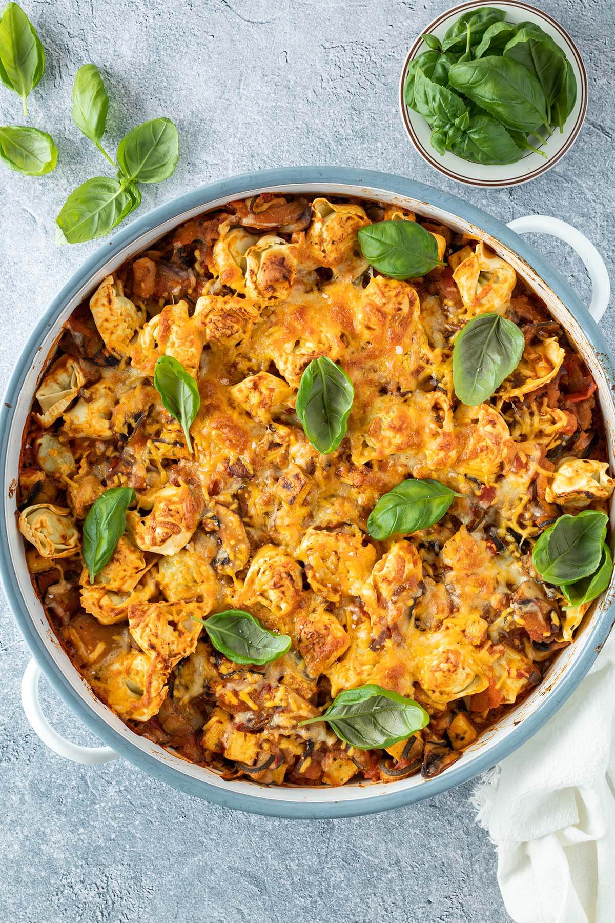 Tortellini Auflauf in einer runden Auflaufform mit frischem Basilikum serviert auf einem hellgrauen Untergrund.