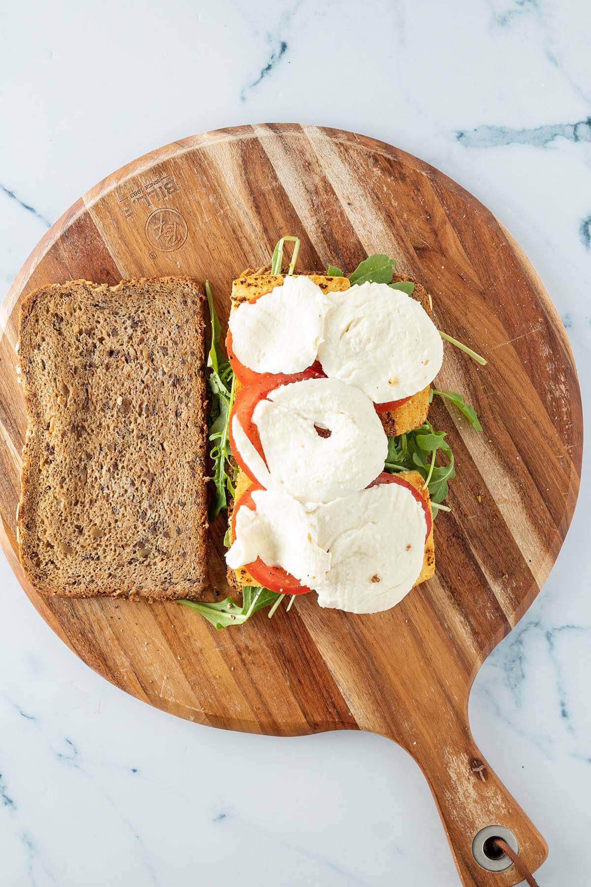 Mit Tofu, Tomate, Mozzarella und Rucola belegtes Brot, daneben eine zweite Brotscheibe.