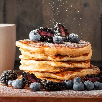 Ein Stapel Vollkorn Pancakes mit Beeren, auf den Puderzucker gestreut wird.