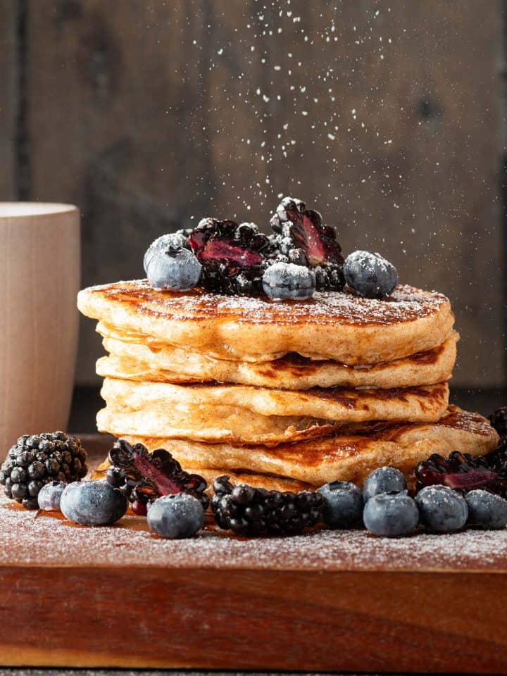 Ein Stapel Vollkorn Pancakes mit Beeren, auf den Puderzucker gestreut wird.