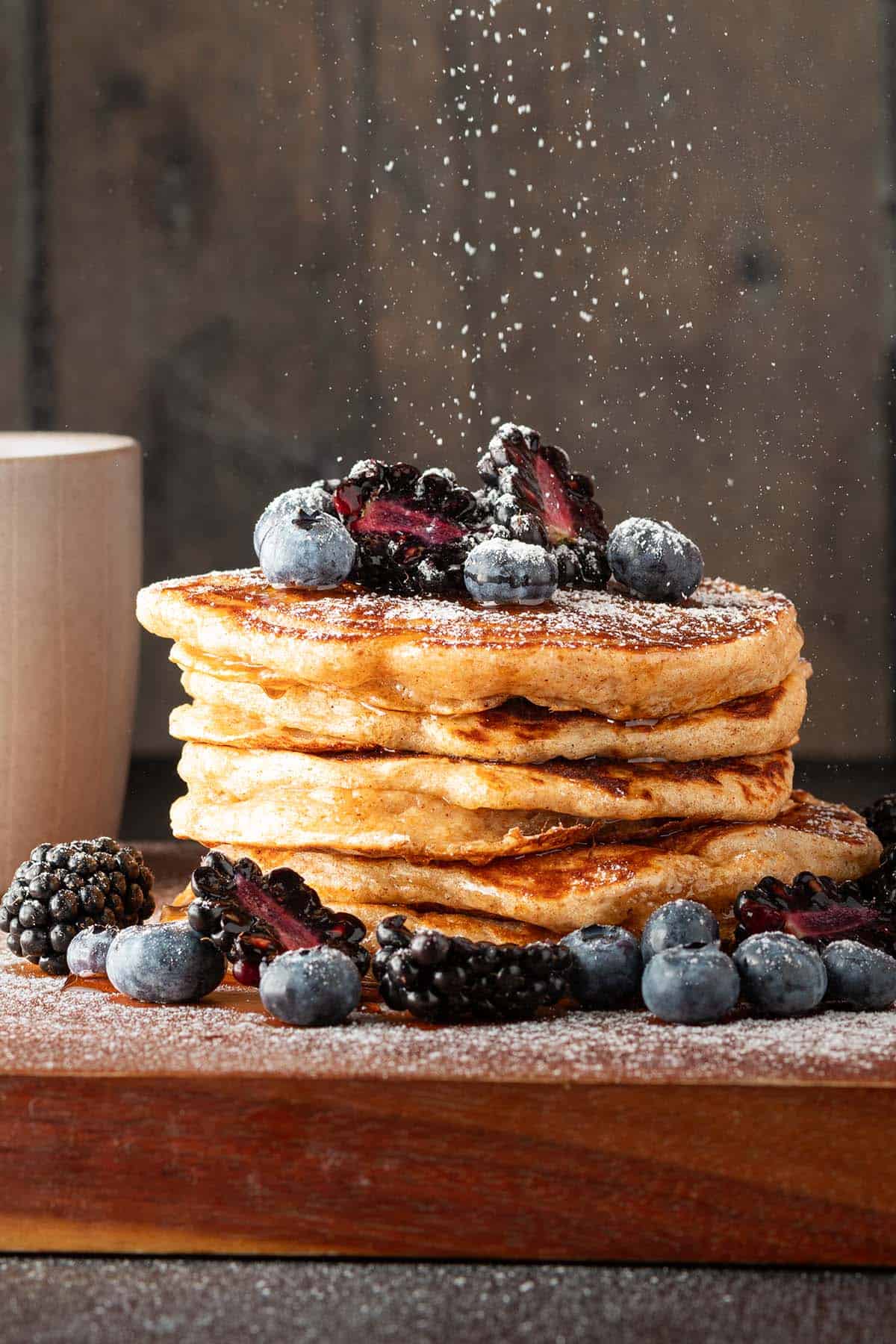 Ein Stapel Vollkorn Pancakes mit Beeren, auf den Puderzucker gestreut wird.