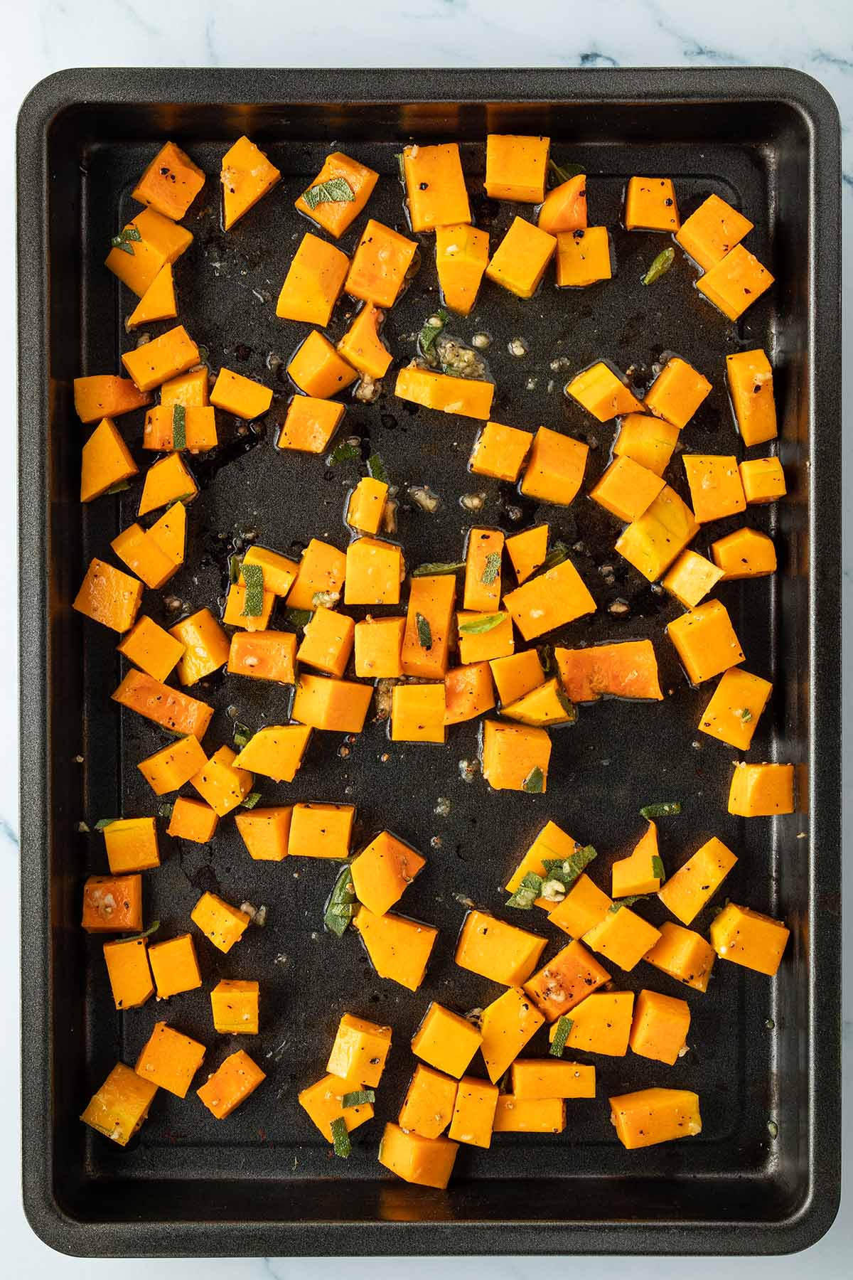 Mit &Ouml;l, Salbei und Knoblauch marinierte Kr&uuml;bisw&uuml;rfel auf einem Blech.
