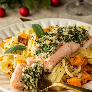 Frontansicht auf ein Teller mit Lachsfilet und Kr&auml;uterkruste auf cremiger Zitronenpasta mit K&uuml;rbis.