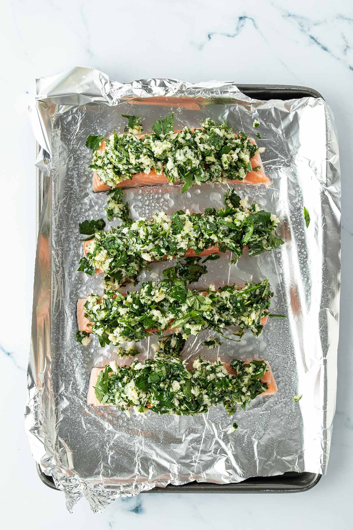 Vier Lachsfilets mit einer Kr&auml;uterkruste auf einem mit Alufolie ausgekleideten Blech.