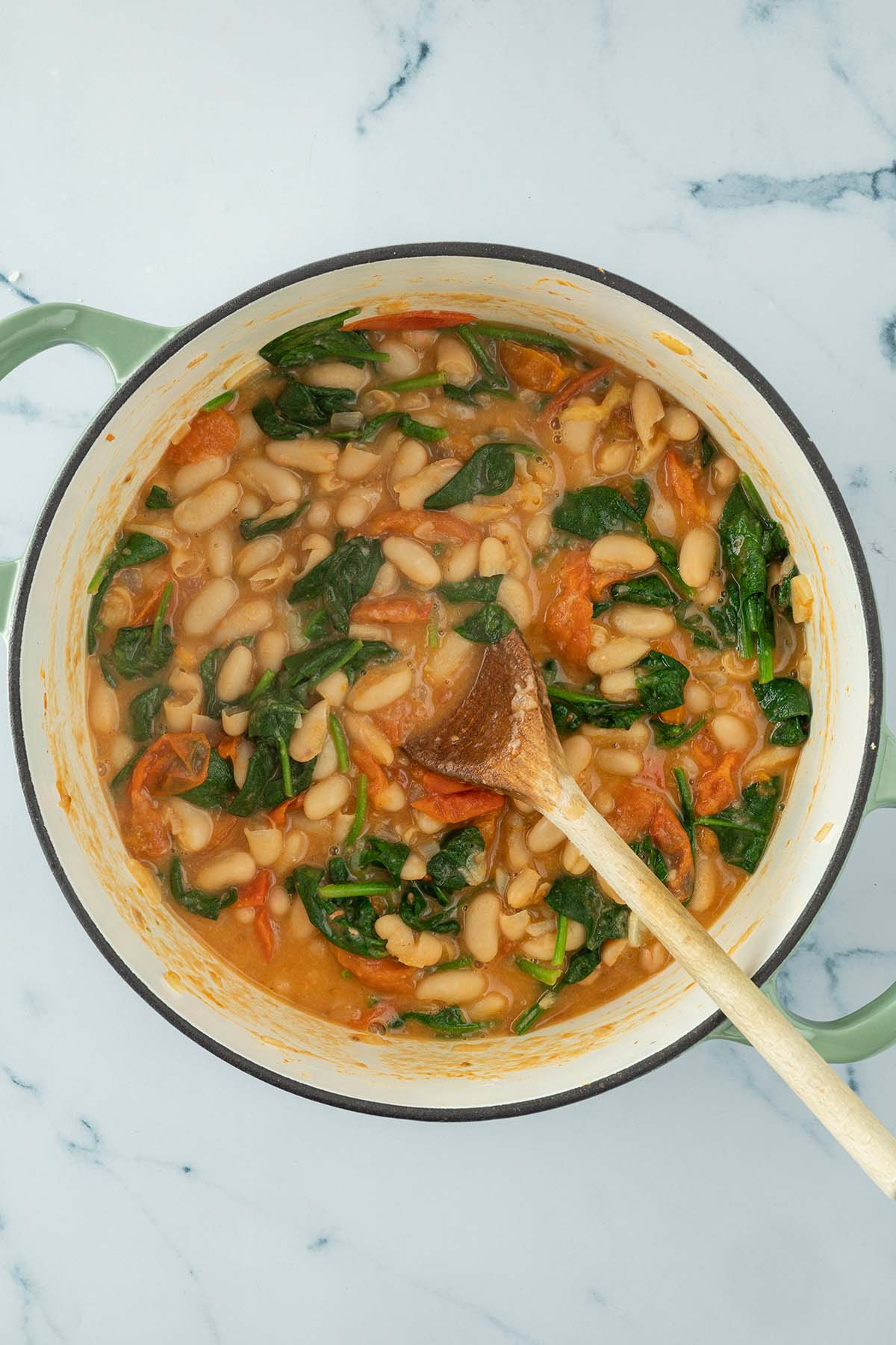 Vogelperspektive auf einen Topf mit wei&szlig;en Bohnen in einer Tomatensauce mit frischem, zusammengefallenen Spinatbl&auml;ttern.