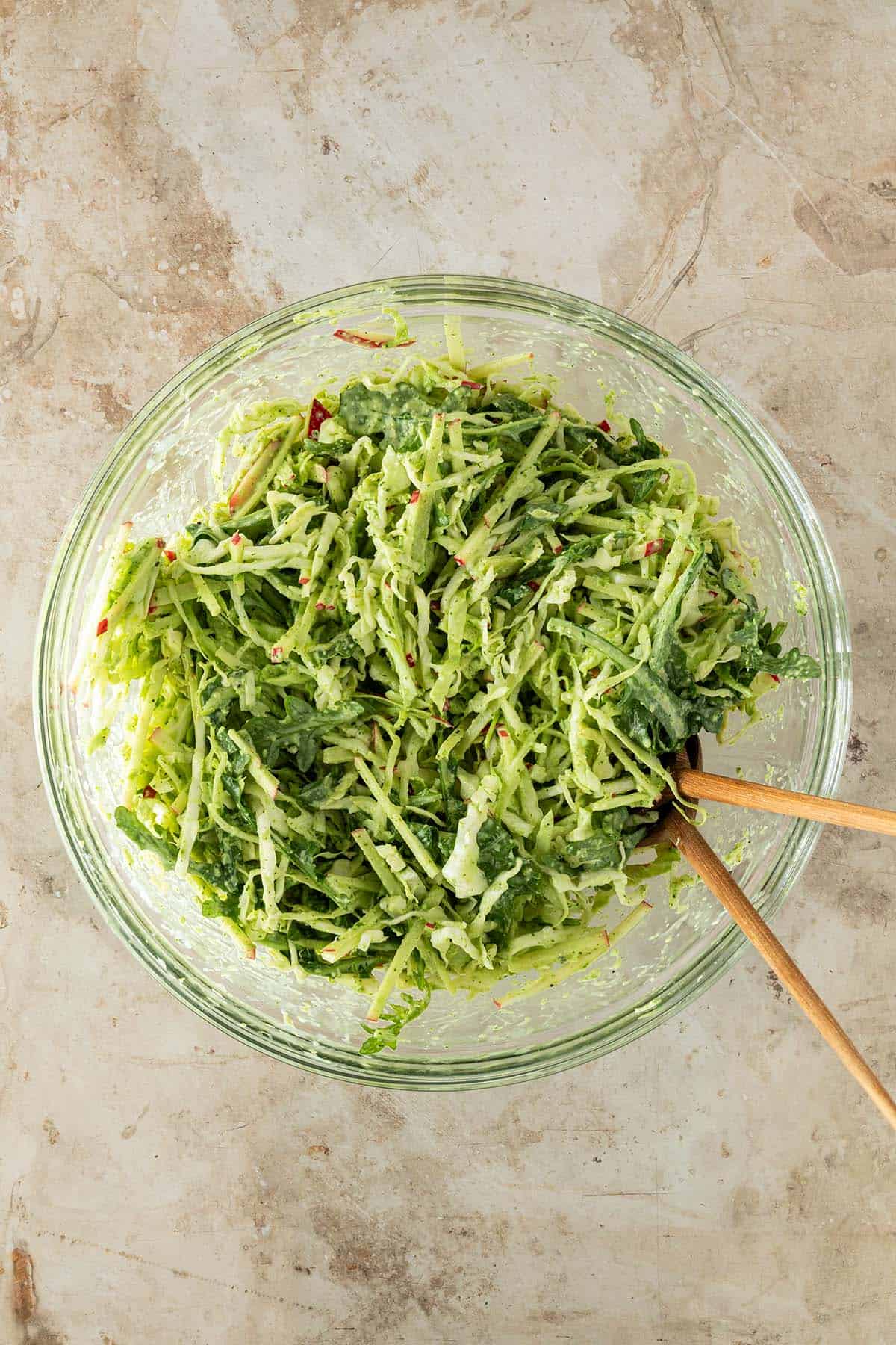 Eine Sch&uuml;ssel mit Wei&szlig;kohlsalat mit Apfel, Rucola und gr&uuml;nem Dressing auf einem Steinuntergrund mit Salatbesteck.