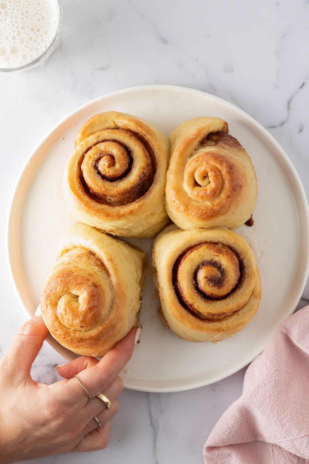 Vier goldbraun gebackene Zimtschnecken auf einem wei&szlig;en Teller, eine Hand l&ouml;st vorsichtig eine Schnecke ab.