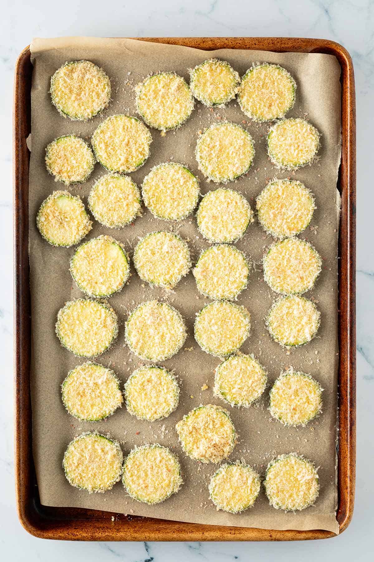 Panierte Zucchini-Scheiben mit Parmesan und Semmelbr&ouml;seln auf einem Backblech.